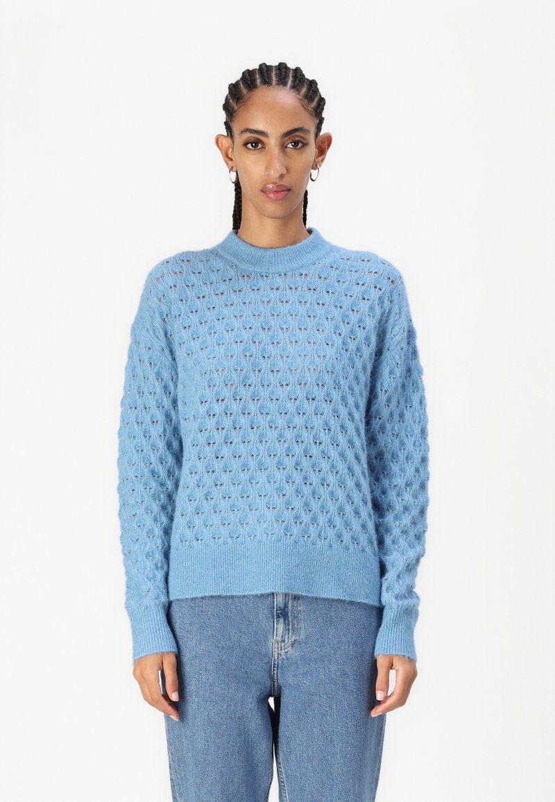 Hellblaues Strickpullover mit strukturiertem Muster, Rundhalsausschnitt und gerippten Bündchen. Kombiniert mit hochgeschnittenen Jeans.