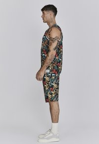 Ujj nélküli virágmintás tank top és hozzáillő fekete rövidnadrág élénk trópusi mintákkal. Fehér kiegészítők és laza fehér edzőcipő jellemzi.