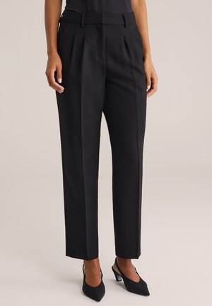 Frau trägt hoch taillierte schwarze, maßgeschneiderte Hosen mit vorderen Falten und schwarze Slingback-Absatzschuhe mit spitzer Spitze vor neutralem Hintergrund.