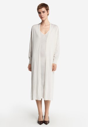 Langer, offener Cardigan in Creme, aus leichtem Stoff gefertigt. Mit überschnittenen Schultern und einer lockeren Passform, kombiniert mit einem schlichten Kleid darunter.