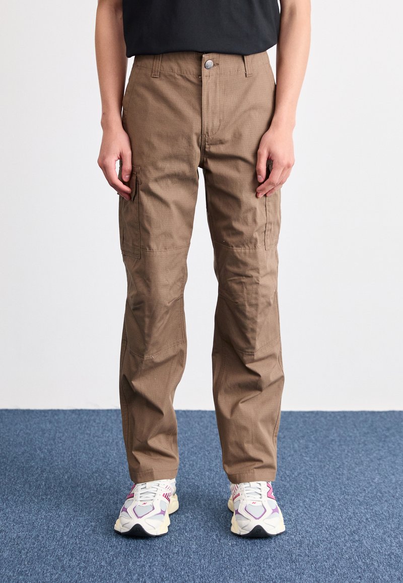 Pantalones cargo marrón claro de corte recto, con bolsillos laterales, pasadores para cinturón y cierre de botón en la parte delantera. Combinados con zapatillas blancas.