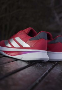 Zapatillas deportivas rojas con parte superior de malla, tres franjas blancas en el lateral, acentos grises y una suela blanca acolchada con detalles en púrpura.