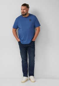 Niebieska koszulka z krótkim rękawem i okrągłym dekoltem, z logo na piersi, w połączeniu z ciemnymi dżinsami i białymi sneakersami na neutralnym tle.