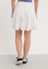 Personne portant une jupe en couches blanche et des baskets noires Adidas avec des rayures blanches, vue de dos sur un fond uni.
