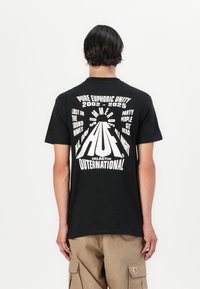 Zwarte katoenen T-shirt met gedurfde witte grafische tekst op de achterkant. De tekst bevat "Zomer van 2002-2025" met een centraal logo-ontwerp.
