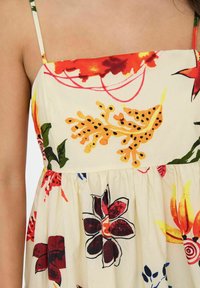 Robe florale en tissu blanc avec des motifs colorés. Présente un décolleté carré, des bretelles fines et une taille froncée. Accents notables en rouge, jaune et vert.