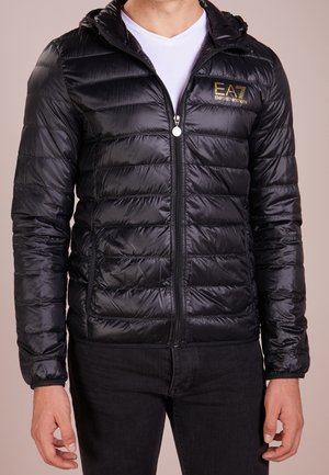 Man wearing black quilted zip-up jacket with "EA7 Emporio Armani" logo on left chest, white t-shirt underneath, and black pants.
