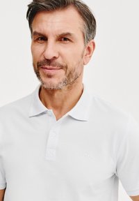 Biała koszulka polo z zapięciem na guziki, z subtelnym wytłoczonym logo i minimalistycznym designem. Tkanina wydaje się gładka i lekka.