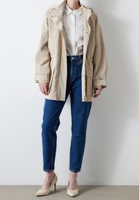 Beżowa, oversize'owa kurtka na zamek, z kieszeniami i regulowanym paskiem w talii, noszona na białej koszuli i niebieskich dżinsach z wysokim stanem, w zestawie z cielistymi szpilkami.