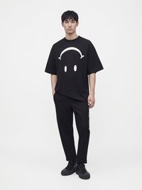 Jeune homme debout, portant un t-shirt noir oversize avec un graphisme minimaliste de visage blanc, un pantalon noir et des baskets noires sur fond blanc.