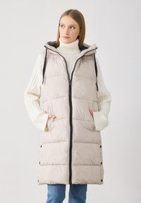 Lett, beige dunjakke uten ermer med hette, frontlås og sidelommer. Har et quiltet design med svarte detaljer langs glidelåsen.