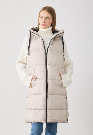 Gilet en duvet léger, beige, avec capuche, fermeture éclair sur le devant et poches latérales. Il présente un design matelassé avec des accents noirs le long de la fermeture éclair.