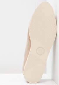 Chaussure en daim beige avec une surface lisse et texturée. La semelle extérieure est en caoutchouc beige clair avec un motif de semelle. Le logo apparaît sur la semelle extérieure.