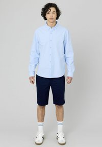 Chemise bleu clair à manches longues, avec un col ; associée à un short bleu marine et des baskets blanches avec des accent noirs et des chaussettes rayées.