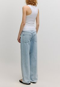 Weißes ärmelloses Oberteil mit Rundhalsausschnitt, kombiniert mit hellblauen weiten Jeans mit Gesäßtaschen. Schlichter Hintergrund.