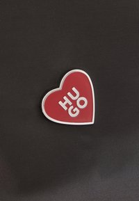 Κόκκινη καρδιακή καρφίτσα με λευκό περίγραμμα και το κείμενο «HUGO» σε λευκά γράμματα πάνω σε υφασμάτινο μαύρο φόντο με υφή.