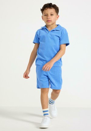 Poika kävelee eteenpäin sininen lyhythihainen kauluspaita yllään, samanväriset shortsit, valkoiset lenkkarit ja valkoiset sukat, joissa on siniset raidat, yksivärisellä taustalla.
