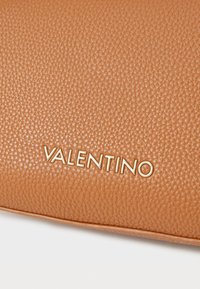 Kumimainen tan-värinen nahkalaukku, jossa on kultainen "Valentino"-logo. Pintarakennetta peittää pikkuinen karheus, mikä tuo esiin pehmeän ja kestävän muotoilun.