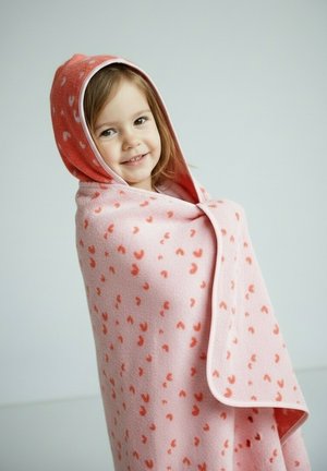 Young girl smiling, wrapped in a pink hooded blanket with small red heart patterns, against a plain light background.