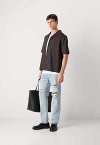 Camisa de manga corta en negro con gran impresión marrón de "HUGO", combinada con unos vaqueros claros desgastados y zapatos negros, sosteniendo una bolsa tote negra.