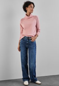 Heller rosa gestrickter Pullover mit hohem Kragen, kombiniert mit weiten blauen Jeans und weißen Sneakern. Der Stoff wirkt weich und strukturiert.