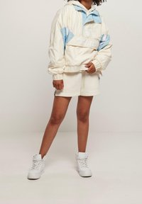 Rompevientos ligero de crema con acentos azules, puños elásticos y capucha, combinado con pantalones cortos beige y zapatillas altas blancas.