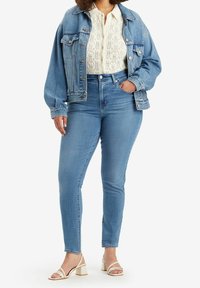 Vaqueros de mezclilla azul claro de cintura alta, combinados con una blusa blanca texturizada y una chaqueta de mezclilla oversized a juego. Sandalias de tacón blancas.