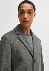 Grau gemischter Wollmantel mit Reverskragen und einem Knopfverschluss, getragen über einem grauen Pullover, mit glatter Textur und tailliertem Schnitt.