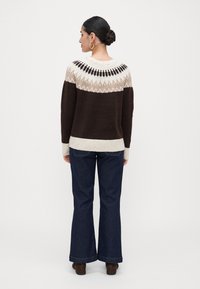 Kvinne med hestehale, iført hoopsmykker, en brun og beige mønstret genser, mørkeblå utsvingte jeans og brune ankelstøvler, vender bort.