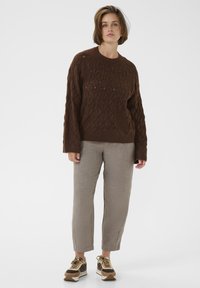 Pull en maille marron avec un motif texturé en losanges, associé à un pantalon en lin beige clair et des baskets marron avec des accents jaunes.