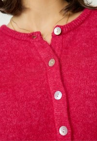 Cárdigan rosa fuerte con un cuello acanalado, que cuenta con seis botones de perla en el frente. Textura suave, de punto, con un ligero brillo.