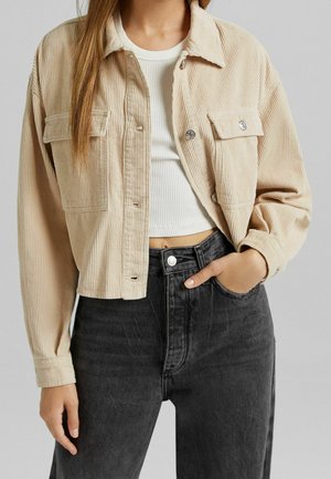 Vrouw draagt een beige cropped corduroy jas over een witte geribde crop top, gecombineerd met zwarte high-waisted jeans, hand in de zak.