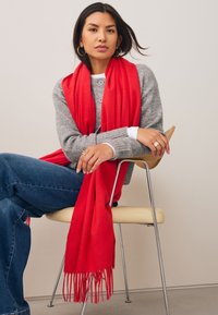 Écharpe rouge aux extrémités à franges drapée sur un pull gris, assortie à un jean bleu, assise sur une chaise à assise claire et lisse.