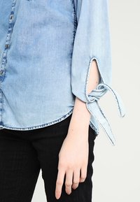 Camicia in denim azzurro chiaro con bottoni, maniche arrotolate con dettagli legati e orlo curvo, abbinata a pantaloni neri a righe.
