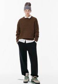Brązowy sweter na białej koszuli z kołnierzykiem, czarne luźne spodnie oraz szare buty z zielonymi sznurówkami. Czapka ma niebieskie i brązowe paski.