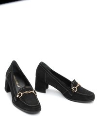 Mocasines de bloque de tacón de ante negro con detalle de cadena dorada y costuras blancas sobre un fondo blanco, en ángulo para mostrar las vistas frontal y lateral.