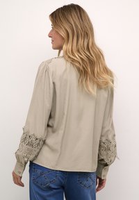 Blouse beige à manches longues avec un bord en dentelle aux manches, dotée d'une coupe décontractée, de poignets à bouton et d'une texture lisse.
