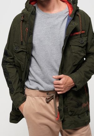 Mann trägt eine olivgrüne Jacke mit orangefarbenem Futter, ein hellgraues T-Shirt und beige Hosen mit Kordelzug, Hand justiert die Vorderseite der Jacke.