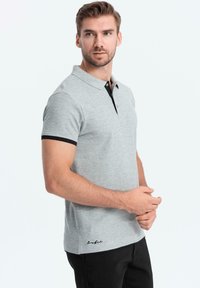 Šedé polo tričko s černým límcem a akcenty na rukávech. Má jemnou strukturu a vyšívané logo poblíž lemu. Úzký střih.