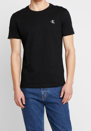 Mężczyzna noszący czarną koszulkę z krótkim rękawem Calvin Klein z logo na piersi i tekstem na ramieniu, w połączeniu z niebieskimi dżinsami.