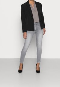Zwarte blazer over een wit met zwart gestreepte blouse, gecombineerd met lichtgrijze skinny jeans en zwarte schoenen met spitse neus tegen een effen achtergrond.