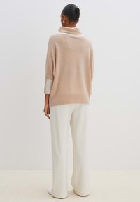 Beige Oversize-Pullover mit hohem Kragen und geripptem Saum, kombiniert mit weißen weit geschnittenen Hosen, die weiche Texturen und eine lässige Passform zeigen.