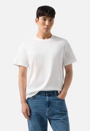 Ung mand med sort hår iført en enkel hvid t-shirt med korte ærmer og blå jeans med den ene hånd i lommen mod en hvid baggrund.