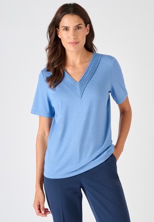 Femme aux longs cheveux bruns portant un t-shirt bleu à manches courtes et col en V avec un détail plissé, ainsi qu'un pantalon bleu marine, debout avec une main dans la poche.