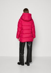 Chaqueta acolchada rosa con capucha, con diseño acolchonado y textura suave. Llevada sobre pantalones negros de cuero sintético y botas negras gruesas.