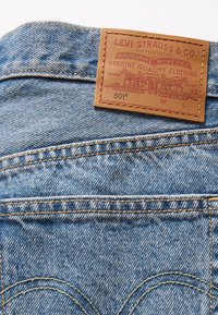 Jean en denim de couleur bleu clair, avec des coutures jaunes contrastantes et une pièce en cuir marron embossée d'un logo et de détails sur la ceinture.