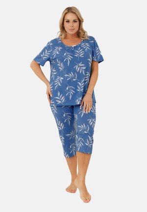 Kobieta stojąca na boso, ubrana w niebieską bluzkę z krótkim rękawem i spodnie 3/4 z białymi liściastymi wzorami na jednolitym tle.