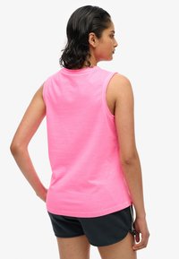 Vrouw met nat, donker haar, gekleed in een felroze mouwloze top en zwarte korte broek, staand met haar rug en rechterzijde zichtbaar.