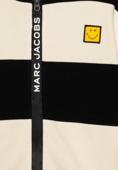 Giacca con zip a strisce in crema, nero e morbida. Presenta una toppa a forma di faccina sorridente gialla e il marchio "MARC JACOBS" sulla zip.