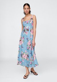 Blå blomsterkjole med rosa og grønne blomster, tettsittende overdel, flagrende midilengde skjørt og brede stropper; kombinert med svarte sandaler.